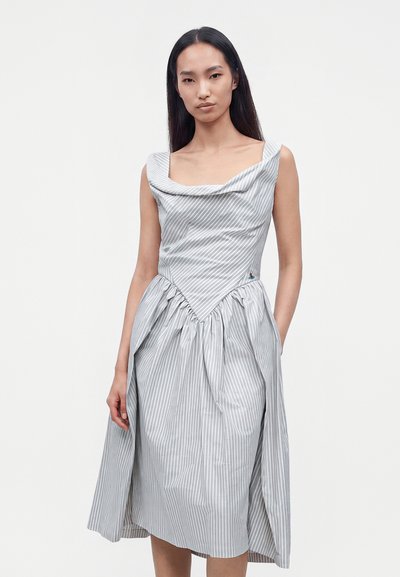 Femme aux longs cheveux noirs portant une robe sans manches rayée gris et blanc avec une jupe froncée et un décolleté asymétrique.
