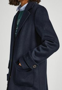 Una persona indossa un soprabito blu scuro con una tasca, sovrapposto a un maglione verde e una camicia blu con colletto, ed è in piedi contro uno sfondo chiaro.
