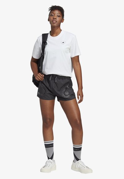T-shirt bianco in cotone, pantaloni corti neri leggeri con vita elastica e tasche, calze a motivo al ginocchio, e sneakers bianche, indossati con uno zaino.