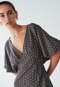 Grå kjole med hvide polkaprikker, dyb V-hals, korte flagrende ærmer og detaljer med rynket talje. Letvægtsstof med glat tekstur.