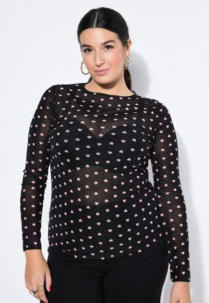 Nainen, jolla on tummat hiukset, musta läpikuultava paita, jossa on vaaleanpunaisia sydämiä, mustat alusvaatteet, hänellä on kultaiset renkaankorvakorut ja vähäinen meikki.