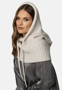Sciarpa con cappuccio lavorata a maglia di colore crema con texture a coste, dotata di lacci. Abbinata a un capo grigio setoso.