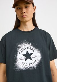 Tamsiai pilkas medvilninis marškinėliai su baltu apvaliu grafiniais elementais, kuriame pavaizduotas juodas žvaigždė ir tekstas "Converse All Star", papildytas purslų efektu.