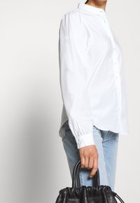 Chemise blanche à boutons avec manches longues, texture lisse, et col. Associée à un denim bleu clair et à un sac à main noir texturé.