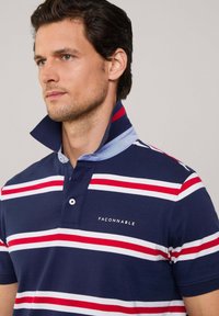 Polo de color azul marino con rayas horizontales rojas y blancas, con un cuello de contraste en azul claro y detalles del logotipo en el pecho.