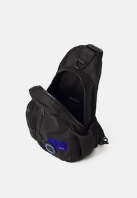 Svart nylon skulderveske med en teksturert overflate, som har et hovedrom med glidelås, et mesh sidelommer og et blått logo-patch. Justerbar skulderstropp inkludert.