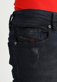 Jeans de mezclilla oscura con un ajuste slim, que presentan un pequeño bolsillo y una etiqueta roja y negra en la parte posterior. Detalles sutiles de desgaste presentes.