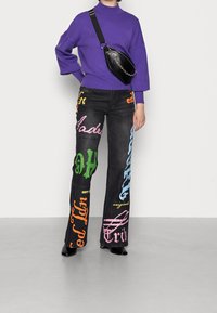 Jeans neri con design testuali colorati in verde, arancione, blu, rosa e viola. Abbinati a un maglione viola e una borsa a tracolla nera.