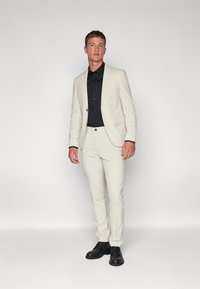 Világos színű testre szabott öltöny fekete inggel és fekete elegáns cipőkkel. Az öltönyön egy gombos zárás található, és slim-fit fazonú.