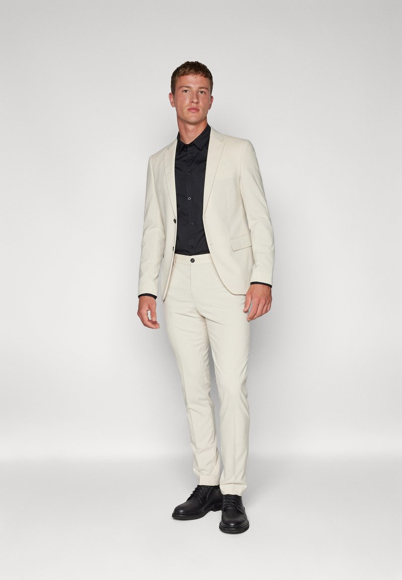 Világos színű testre szabott öltöny fekete inggel és fekete elegáns cipőkkel. Az öltönyön egy gombos zárás található, és slim-fit fazonú.