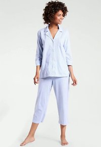 Nainen, jolla on kihara hius, seisoo paljain jaloin päällään vaaleansinisestä raidallisesta napillisesta pyjamapuserosta ja lyhyistä pyjamahousuista, hymyillen ja katsoen oikealle.