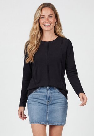 Smilende kvinde med langt blond hår iført hoop-øreringe, en sort langærmet top og en blå denimhjørt mod en ensfarvet baggrund.