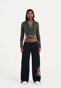 Olijf groene cropped ritssluiting trui met ribbeltextuur, gecombineerd met zwarte cargo broek met zijzakken en kleurrijke geborduurde details.