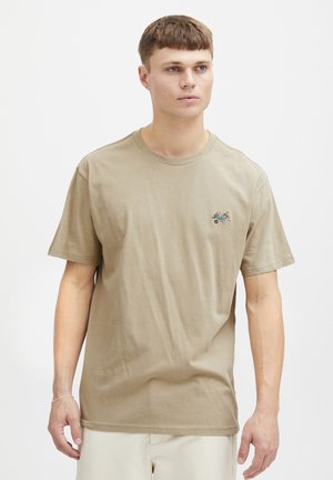 Beige puuvillapaita, jossa on pyöreä kaula-aukko, lyhyet hihat ja pieni brodeerattu kuvio vasemmassa rinnassa. Silkkinen rakenne, rento istuvuus.