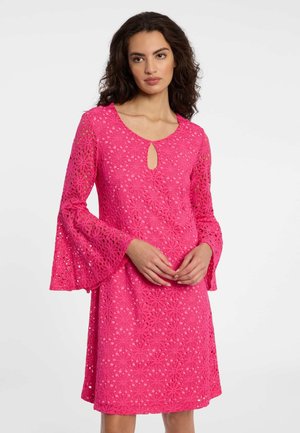 Frau in einem hellpinkfarbenen Spitzenkleid mit Blumenmustern, Glockenärmeln und kleinem Schlitz am Ausschnitt, steht vor einem schlichten Hintergrund.