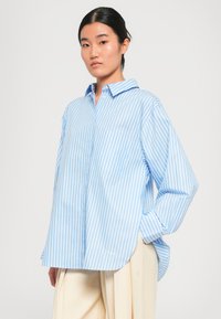 Light blue and white striped oversized shirt with a buttoned collar, front pocket, and relaxed sleeves, made from a smooth fabric.
