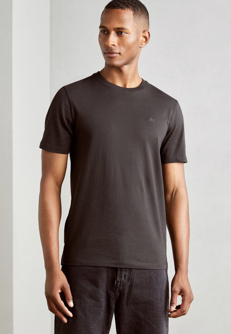 Sorte bomulds T-shirt med korte ærmer, rund halsudskæring og diskret broderet logo på brystet; båret med mørke jeans.