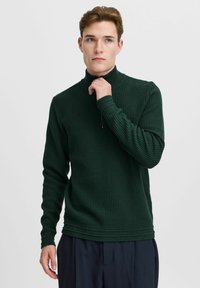 Dunkelgrüner gestrickter Pullover mit hohem Kragen und halbem Reißverschluss. Strukturierter Stoff und eng anliegende Bündchen. Kombiniert mit dunklen Hosen.