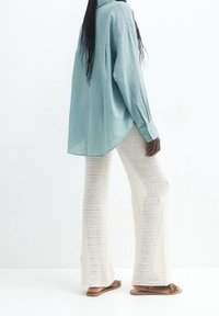 Chemise ample à manches longues de couleur bleu clair en tissu texturé, associée à un pantalon évasé en crochet blanc. Des sandales plates marron complètent le look.