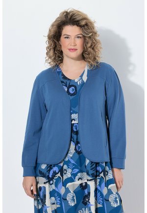 Frau mit lockigem Haar trägt ein blaues Kleid mit Blumenmuster und eine einfarbige, offene blaue Strickjacke und steht vor hellem Hintergrund.