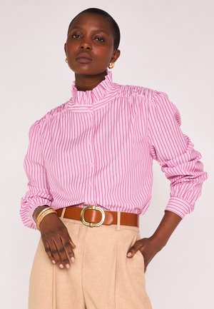 Donna con capelli corti che indossa una blusa rosa a righe con volant, pantaloni beige a vita alta, cintura marrone con fibbia dorata e braccialetti ed orecchini dorati.