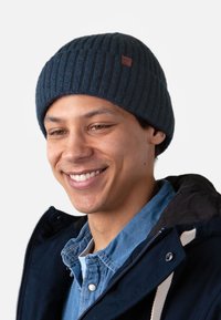Beanie lavorato a maglia di un blu navy scuro con texture a coste, caratterizzato da un piccolo etichetta in pelle. Chi lo indossa è vestito con una giacca scura e una camicia azzurra chiara.