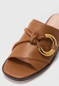 Brune lær slidesandal med knytt strap detalj og gylden sirkulær hardware aksent. Åpen tå design med flat såle.