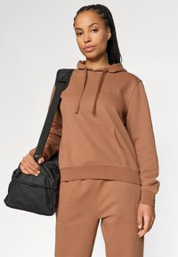 Brun hette laget av mykt stoff, med en frontlomme, snor og en subtil logo. Svart bag med nettdetaljer holdt i modellens hånd.