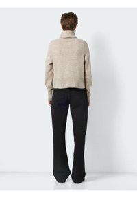 Personne aux cheveux courts portant un pull à col roulé beige et un jean noir droit, debout face à un fond gris clair uni.