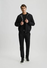 Schwarze Zip-Up-Hoodie mit grauem Futter, kombiniert mit einem schwarzen Rundhalsausschnittshirt, schwarzen Hosen und schwarzen Sneakern mit weißen Sohlen.