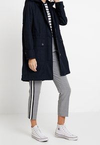 Parka navy con cappuccio, chiusura frontale con cerniera e bottoni, dotata di due tasche laterali e polsini. Indossata con pantaloni corti a quadri e sneakers bianche.