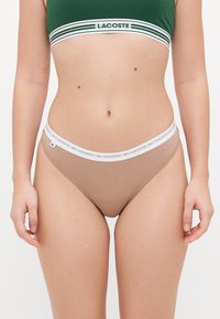 Lacoste rudos maudymosi kelnaitės su baltu prekinio ženklo juosmeniu, pasižyminčios lygiu paviršiumi ir aukštu kirpimu.