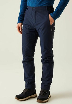 Mężczyzna ubrany w granatowe spodnie outdoorowe z kieszeniami na zamki oraz czarne buty turystyczne, stojący na jednolitym tle.