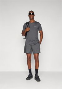 Grå tränings-t-shirt och matchande shorts av lättviktsmaterial. Svart ryggsäck, solglasögon och svarta skor kompletterar outfiten.