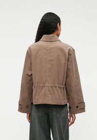 Chaqueta de pana marrón con un corte cropped, gran cuello, cintura elástica y puños abotonados. Lleva puesta sobre pantalones de mezclilla grises.