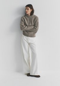 Lichtbruine gebreide trui met dikke kabelpatronen, hoge kraag; gecombineerd met brede witte broek en donkere schoenen, afgebeeld tegen een effen achtergrond.