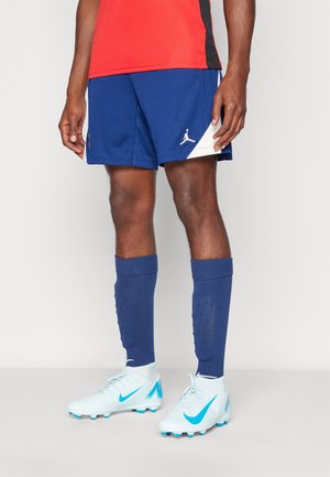 Un footballeur présente un short bleu et des chaussettes montantes jusqu’aux genoux, avec des chaussures de football blanches et un maillot d’entraînement rouge.
