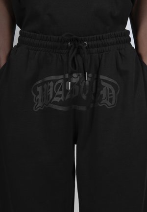 Pantalon à taille élastique noire avec cordon de serrage et logo "WASTED" gris foncé subtil à l'avant, mains reposant dans les poches.