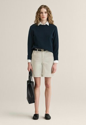 Kvinde med krøllet hår iført en marineblå sweater, hvid skjorte med krave, beige shorts, sorte loafers og holder en sort tote-taske, står mod en ensfarvet baggrund.
