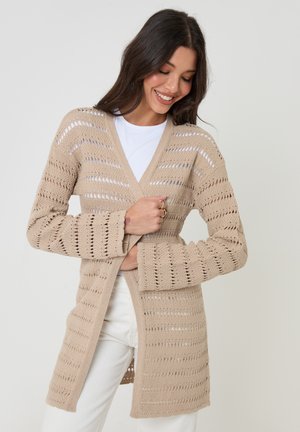 Donna che indossa un cardigan beige a maglia aperta sopra una camicia bianca e pantaloni, sorridente mentre sistema la parte frontale del cardigan.