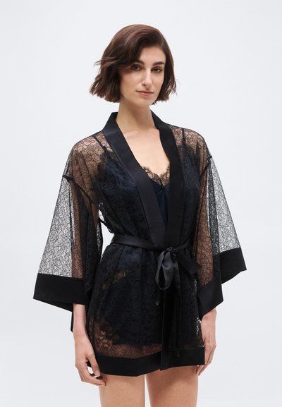 Mujer vistiendo una bata negra de encaje transparente con ribete y cinturón de satén, posando sobre un fondo blanco liso.