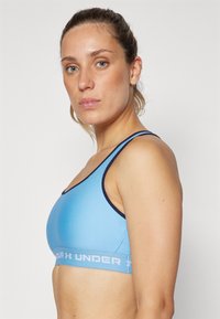 Lyseblå sports-bh med marineblå kant, laget av elastisk materiale; har et logo-bånd for støtte og et enkelt, buet design.