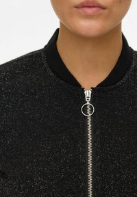 Veste bomber noire avec fermeture à zip et tirette ronde, dotée d'une texture scintillante et d'un col côtelé. Vue en gros plan mettant l'accent sur le décolleté.
