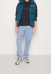 Türkiser Steppweste über einem schwarzen Hoodie, kombiniert mit hellblauen Jeans und mehrfarbigen Sportschuhen. Glatte Texturen, lässige Passform.