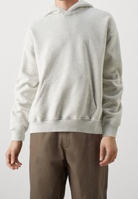 Jasnoszara bluza z kapturem wykonana z miękkiego materiału, z kieszenią kangurką, ściągaczami przy rękawach i luźnym krojem. Noszona z ciemnobrązowymi spodniami.