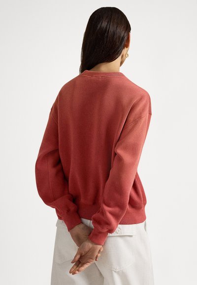 Rdeč bombažni pulover z okroglim vratnim izrezom, dolgimi rokavi in rebrastimi manšetami, nošen obrnjeno z zadržanimi rokami za hrbtom.