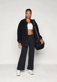 Melnā flīsa jaka ar rāvējslēdzēju, balts apgriezts krekls, melnas bikses un baltas sporta kurpes. Modelis tur melnu plecu somu.