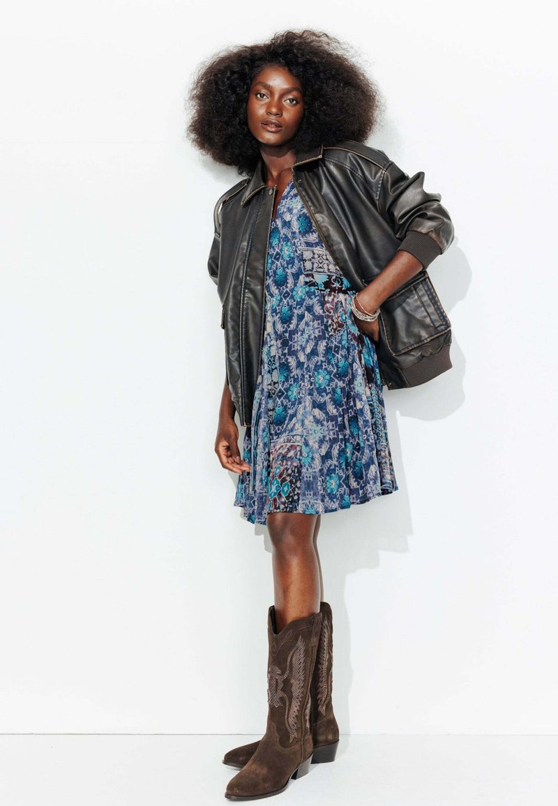 Veste bomber en cuir marron sur une robe à motifs bleus. Bottes de cowboy en suède marron avec des accents embossés. Textures lisses partout.