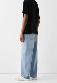 Henkilö, jolla on päällään vaaleansiniset levenevät farkut, musta t-paita ja valkoiset lenkkarit, seisoo tasaisella valkoisella taustalla.