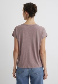 Kurzärmliges, mauvefarbenes Shirt aus weichem Stoff mit feiner Textur. Rundhalsausschnitt und lockere Passform. Kombiniert mit hellblauen Jeans.